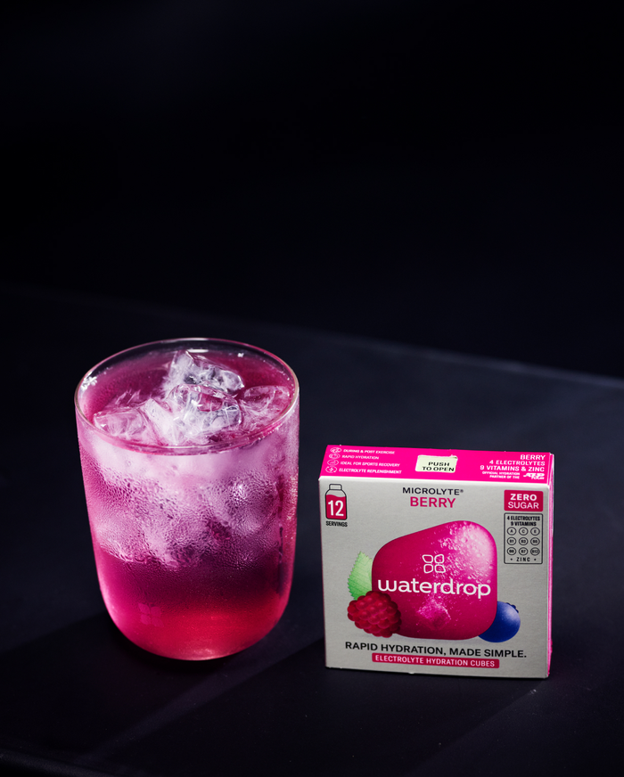 Glass of pink electrolyte drink with ice next to a 12-pack of Microlyte BERRY waterdrop, providing essential electrolytes for sports activities.