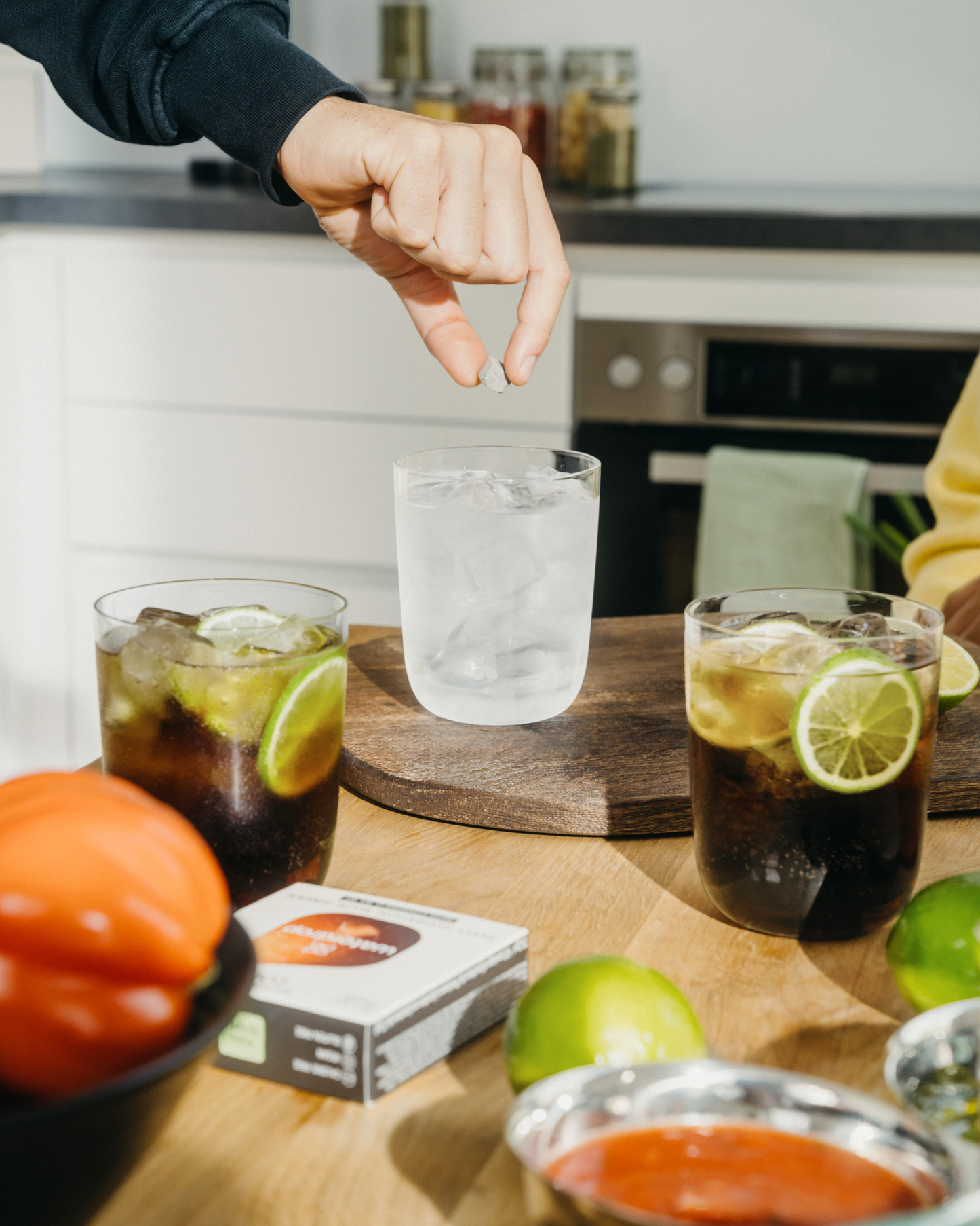 Hand dropping a COLA pill into a glass of ice water, highlighting a sugar-free, calorie-free drink with added vitamins. Limited Edition.