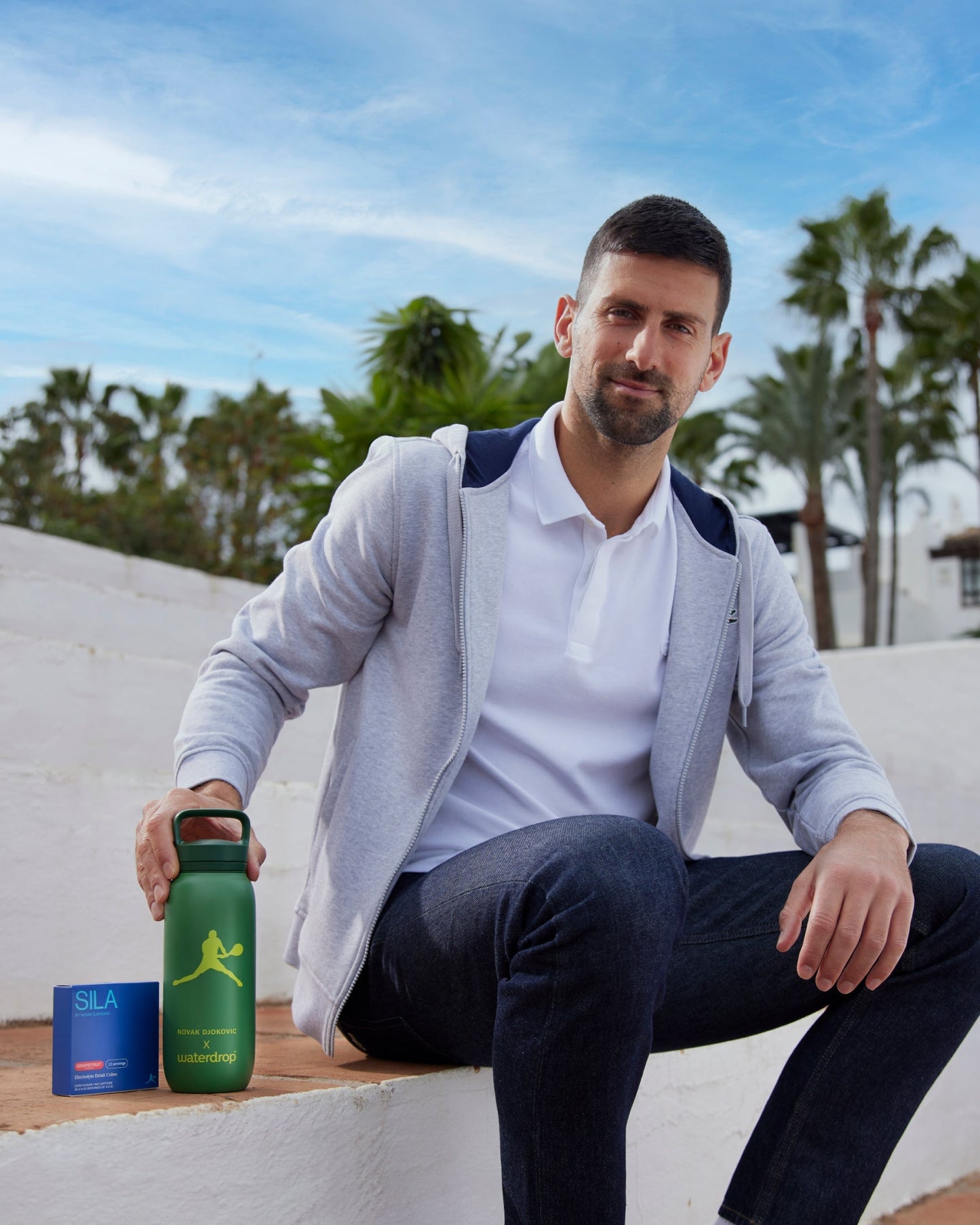 Novak sitting on a bench holding a Novak Djokovic Bottle, featuring a green bottle and a 12 Pack from waterdrop's SILA electrolyte hydration collection.