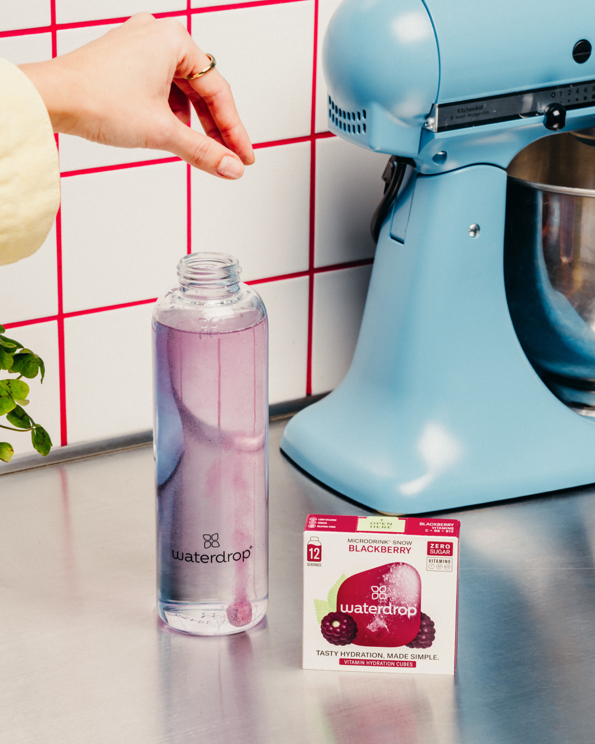 Hand dropping a cube of waterdrop BLACKBERRY Microdrink, with a 12-pack of waterdrop cubes and a glass of liquid nearby.