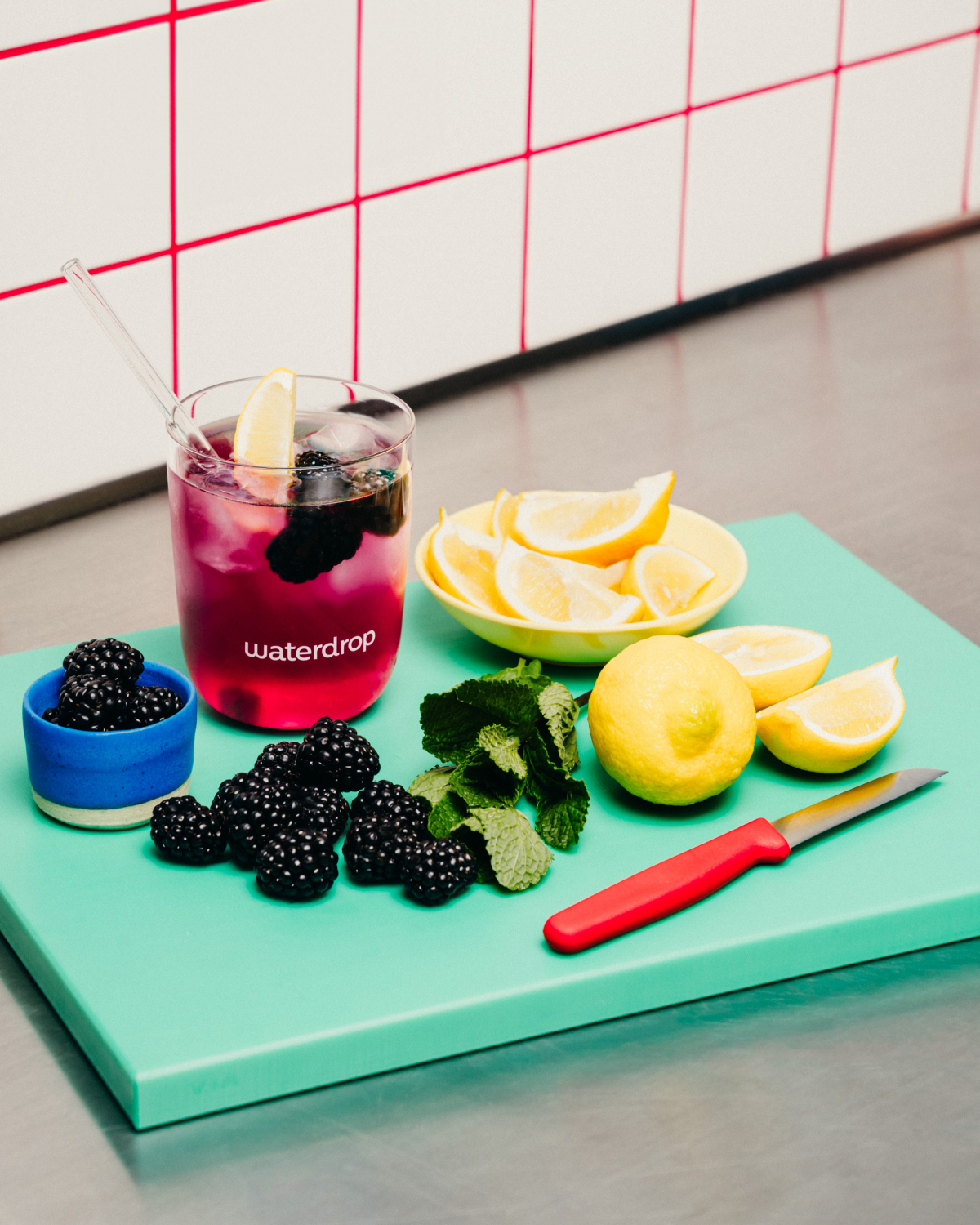 Cutting board with a glass of BLACKBERRY Microdrink, lemons, a knife, a bowl of blackberries, and a bowl of lemons.