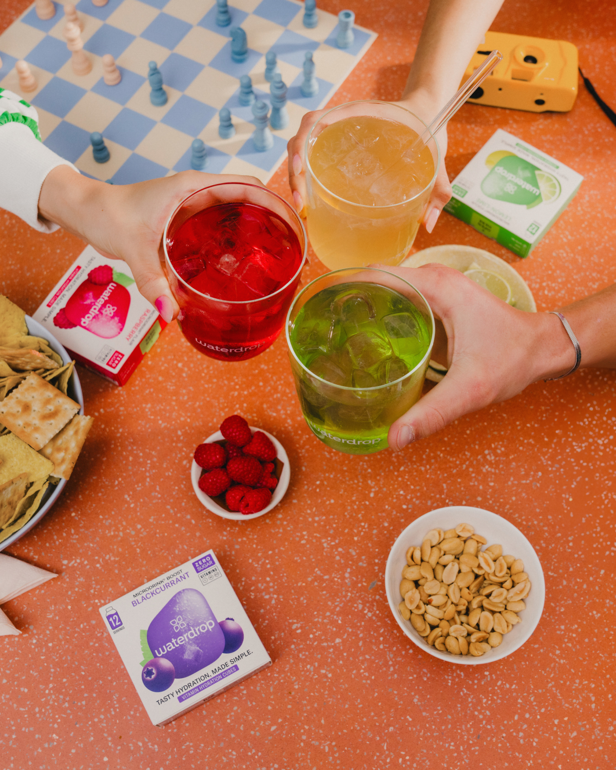Hands holding drinks with ice and snacks, featuring a box of Waterdrop® BLACKCURRANT Microdrink cubes on a table.