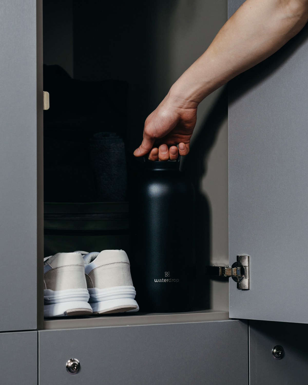 Hand holding a black All-Purpose Thermo Bottle with Loop Lid, with white shoes visible in a cupboard in the background.