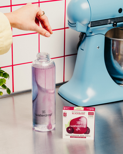 Hand dropping a cube of waterdrop BLACKBERRY Microdrink, with a 12-pack of waterdrop cubes and a glass of liquid nearby.	